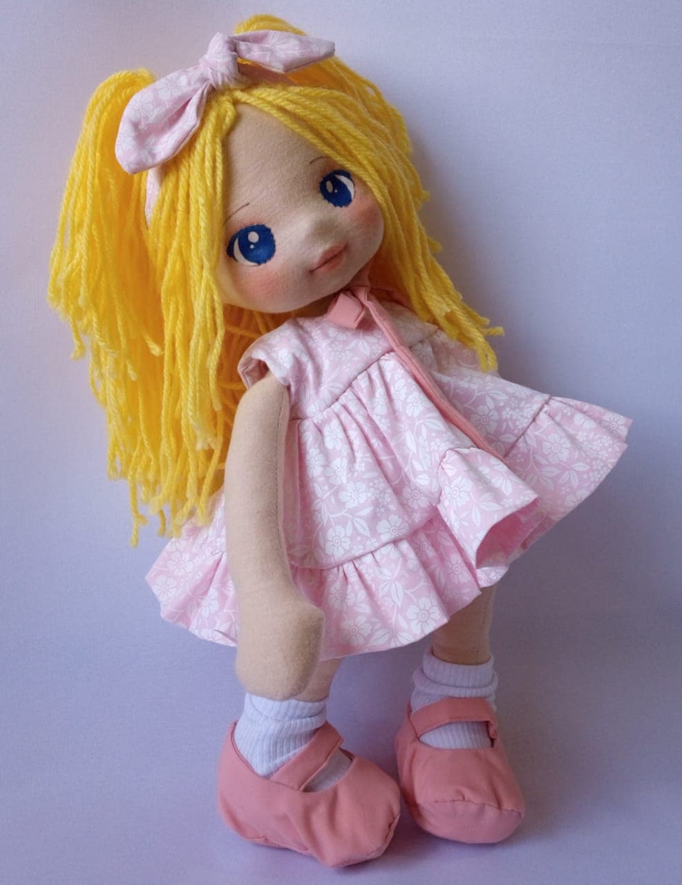 Boneca de pano loira com vestido rosa claro, sapatos rosa escuro e olhos azuis.