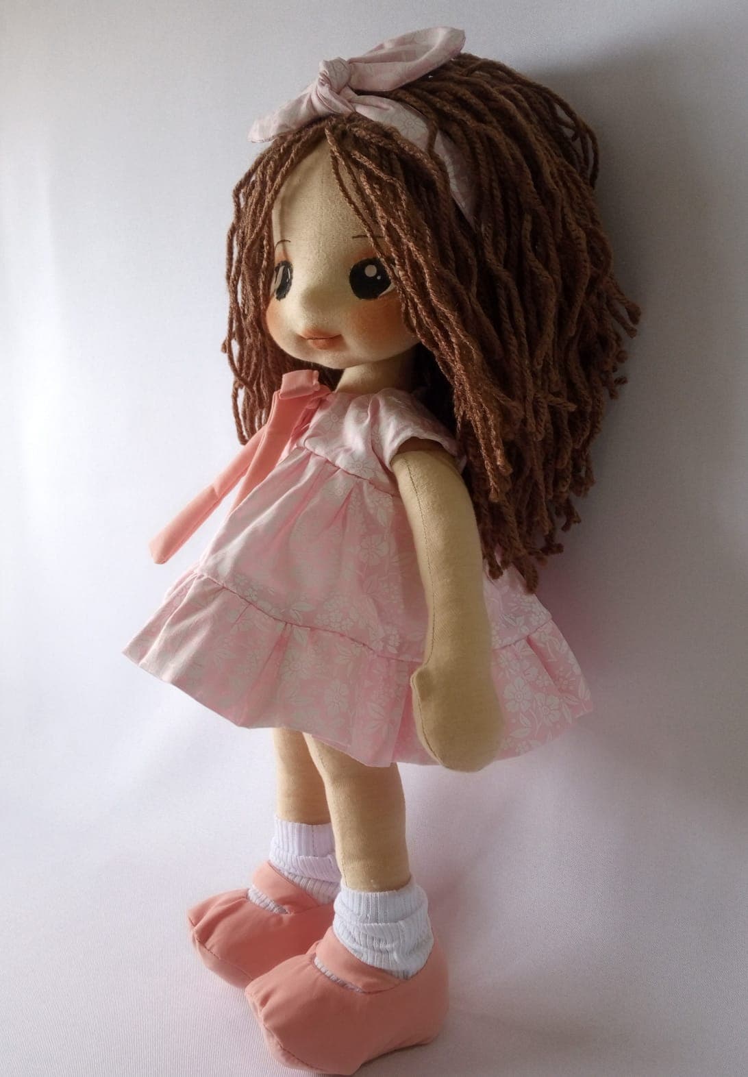 Boneca de pano morena com vestido rosa claro, sapatos rosa escuro e cabelo castanho.