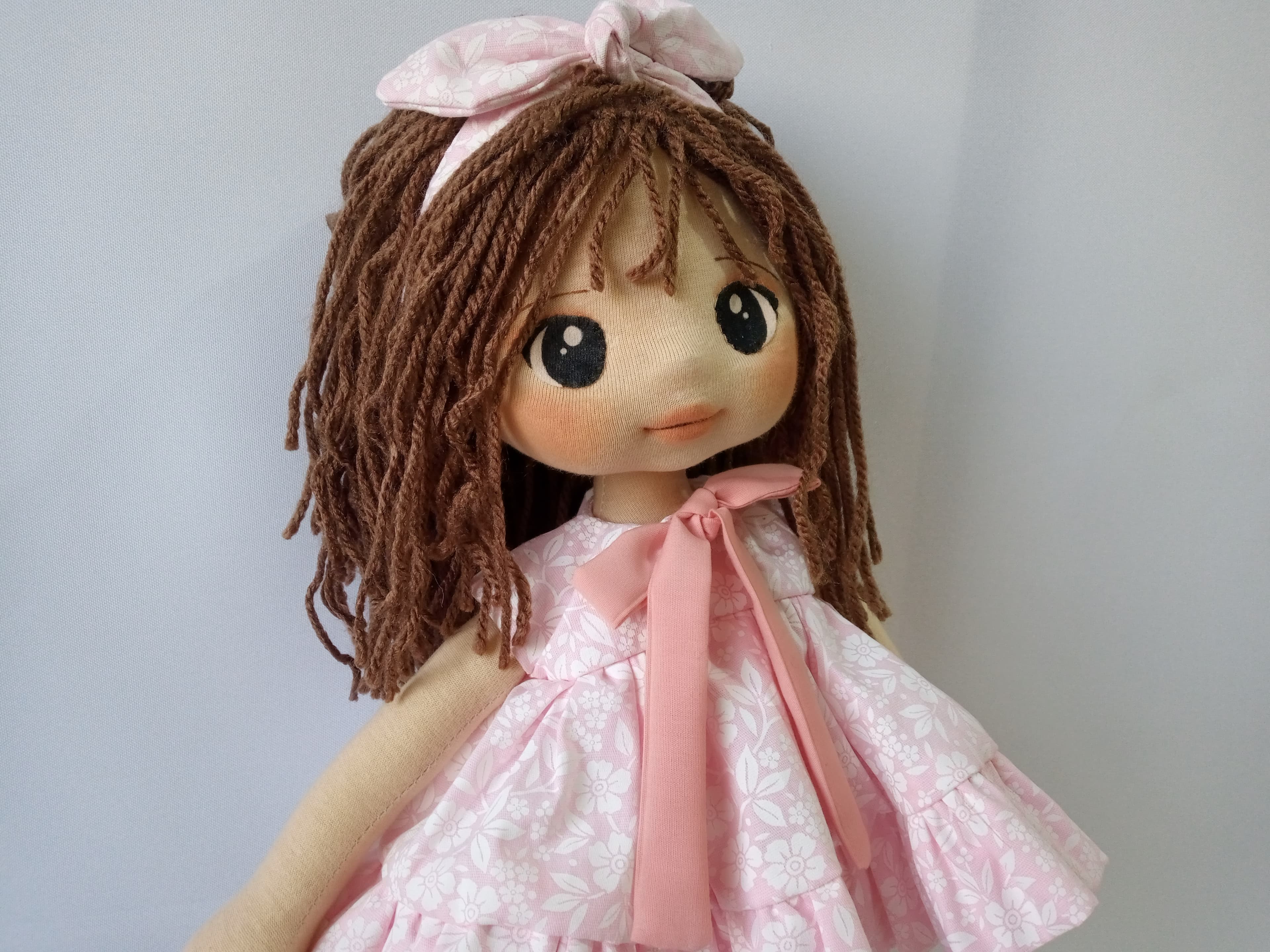 Selfe de boneca de pano morena com vestido rosa claro e cabelo castanho.