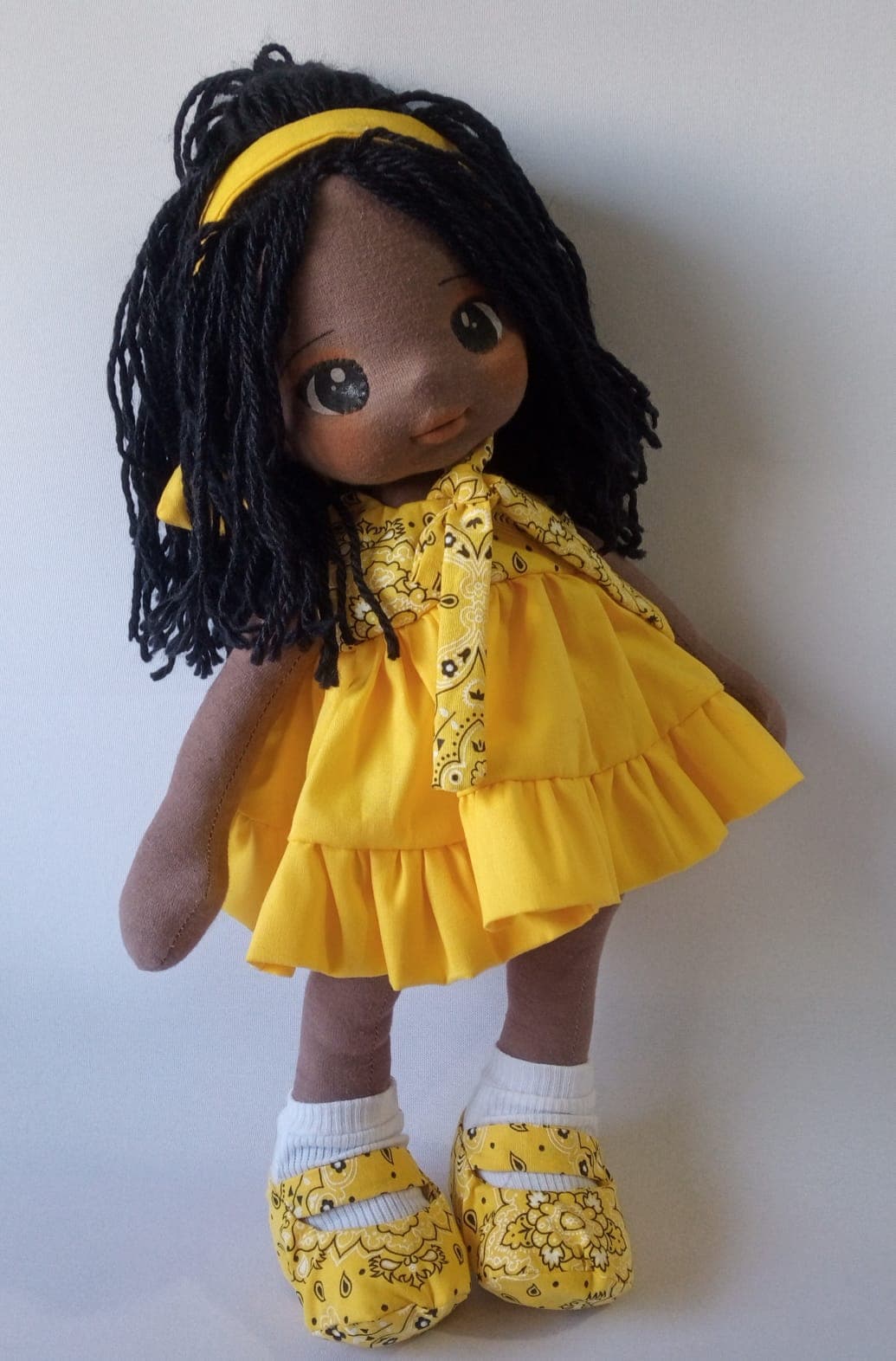 Boneca de pano negra com vestido amarelo e sapatos amarelos, cabelo preto.