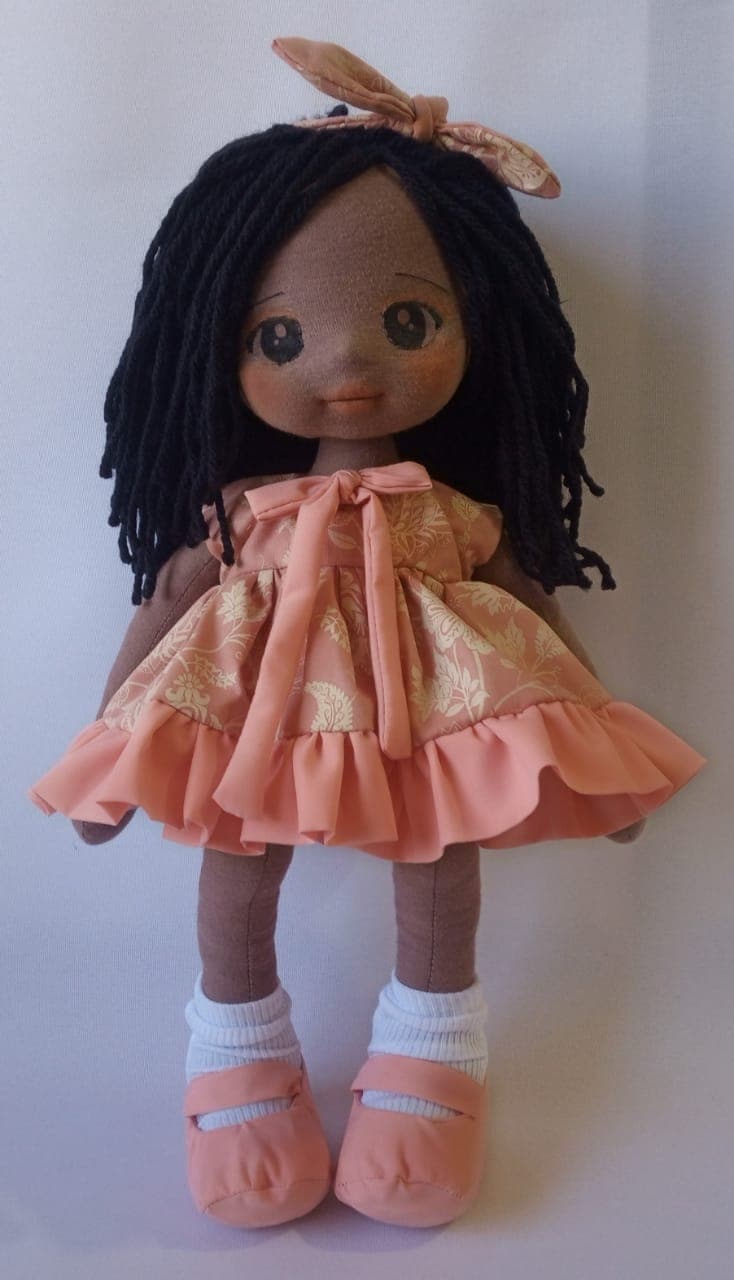 Boneca de pano negra com vestido salmão, flores e sapatos salmão, cabelo preto.