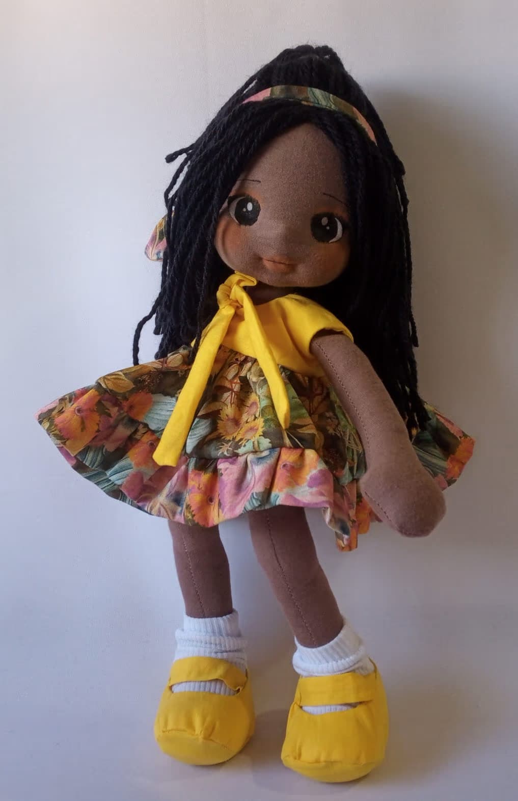 Boneca de pano negra com vestido florido alegre e sapatos amarelos, cabelo preto