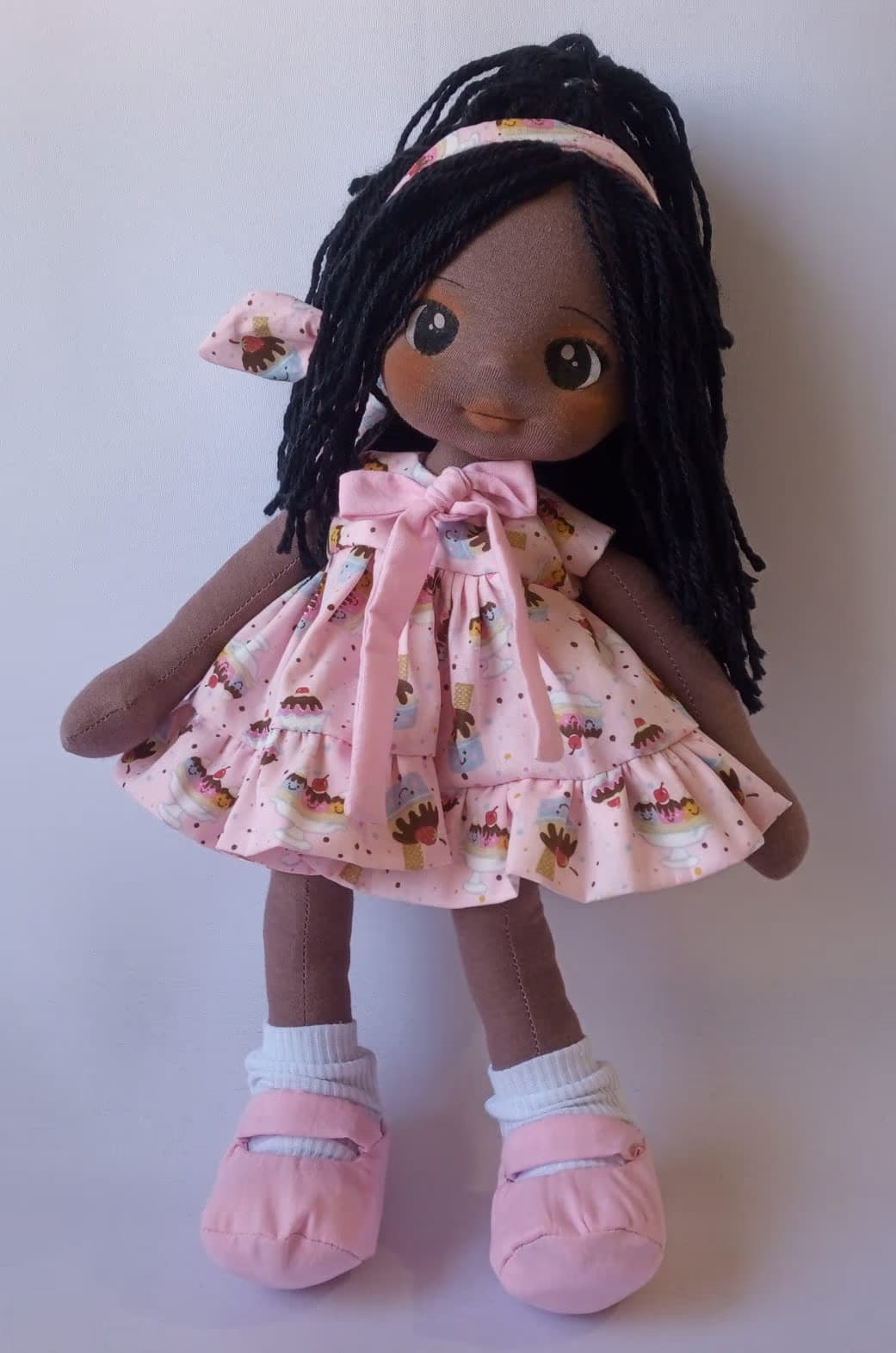 Boneca de pano negra com vestido rosa de docinhos, sapatos rosa e cabelo preto