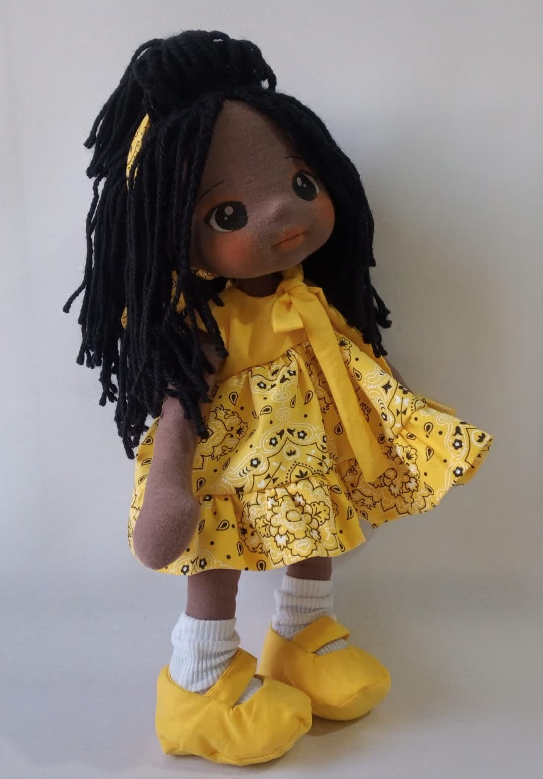 Boneca de pano negra com vestido amarelo, sapatos amarelos e cabelo preso, preto
